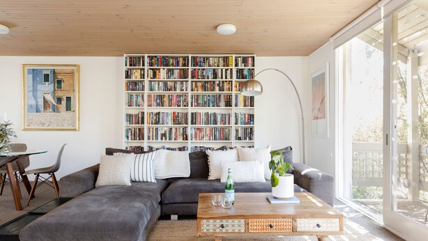 Ein modernes Wohnzimmer mit einer großen Bücherwand, einem grauen Ecksofa, einem Holztisch und einem großen Fenster, das natürliches Licht hereinlässt.
