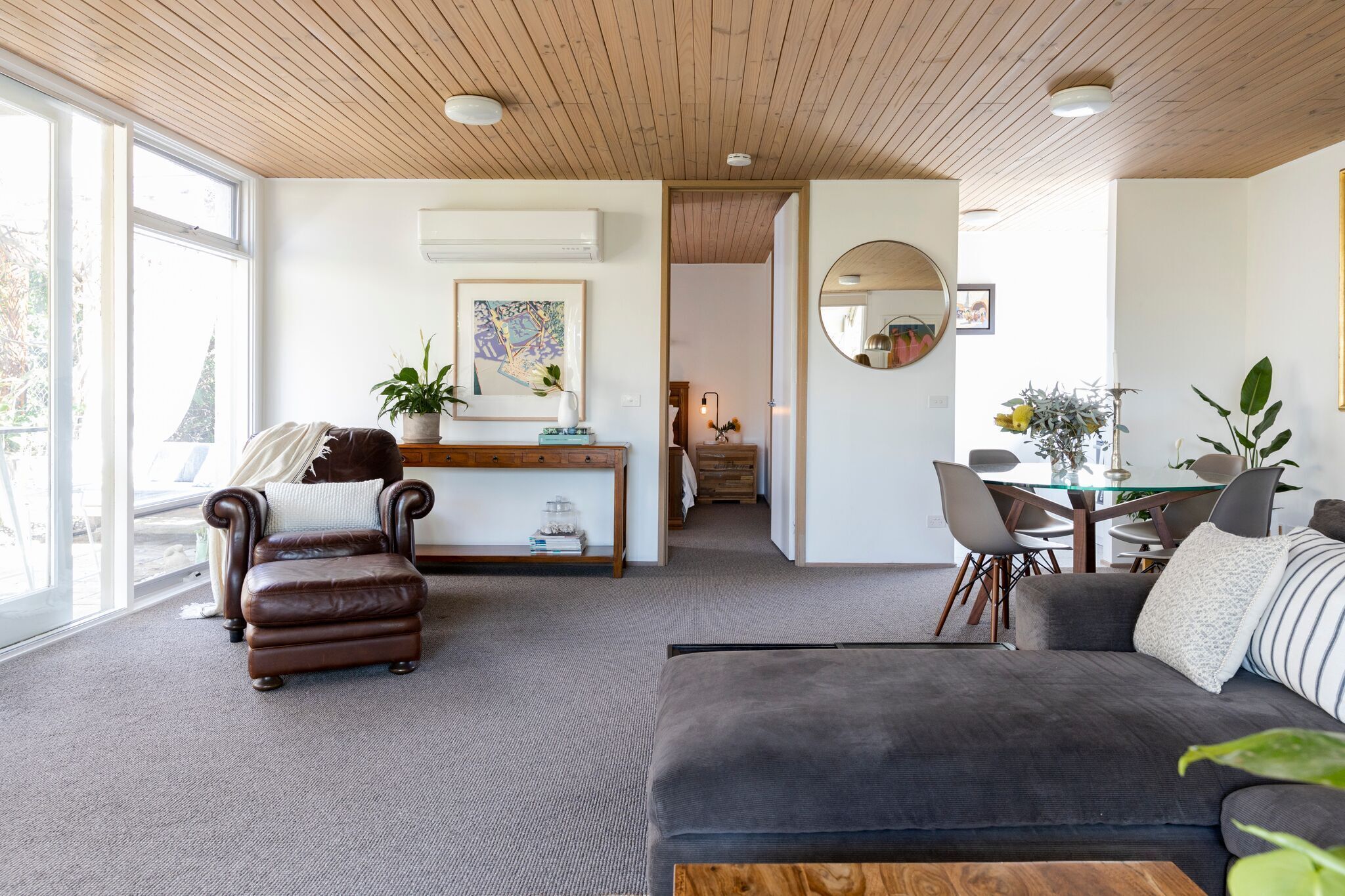 A modern living room with a leather armchair, a grey sofa, a wooden ceiling, and a dining area with a glass table and chairs.