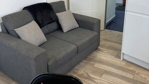 Un sofá gris con varios cojines, una mesa de comedor de madera con un jarrón de flores rosas y una puerta blanca al fondo.