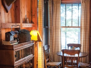 A cozy room with a wooden interior, a table with chairs, a lamp, and a window with curtains. - Signature Cabin 3 with Lake View (Paul Smiths)