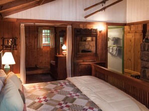 A cozy wooden cabin bedroom with a quilted bed, a wooden dresser, a mirror, and a stone wall. - Signature Cabin 3 with Lake View (Paul Smiths)
