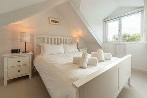 A bedroom with a sloped ceiling, a white bed with pillows and rolled towels, a bedside table with a lamp, and a window with a view of greenery.