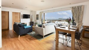 A spacious living area with a wooden floor, a sofa, a dining table with chairs, and a large window offering a view of the sea. - A stunning, environmentally friendly holiday haven at Mor Cliff, in an AONB. (Chapel Porth)