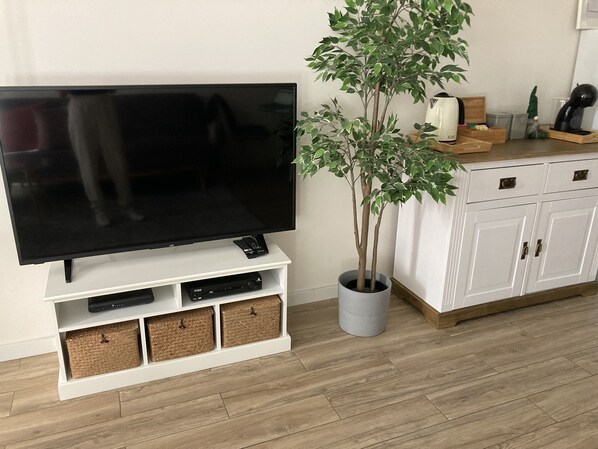 A flat-screen TV on a white stand with storage baskets, a potted plant, and a white cabinet with drawers. - CASA JOY Vilamoura Apartment (Vilamoura)