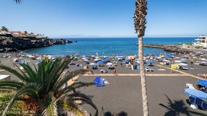 Een strand met talloze parasols en ligbedden, een palmboom en rotsachtige kliffen op de achtergrond.