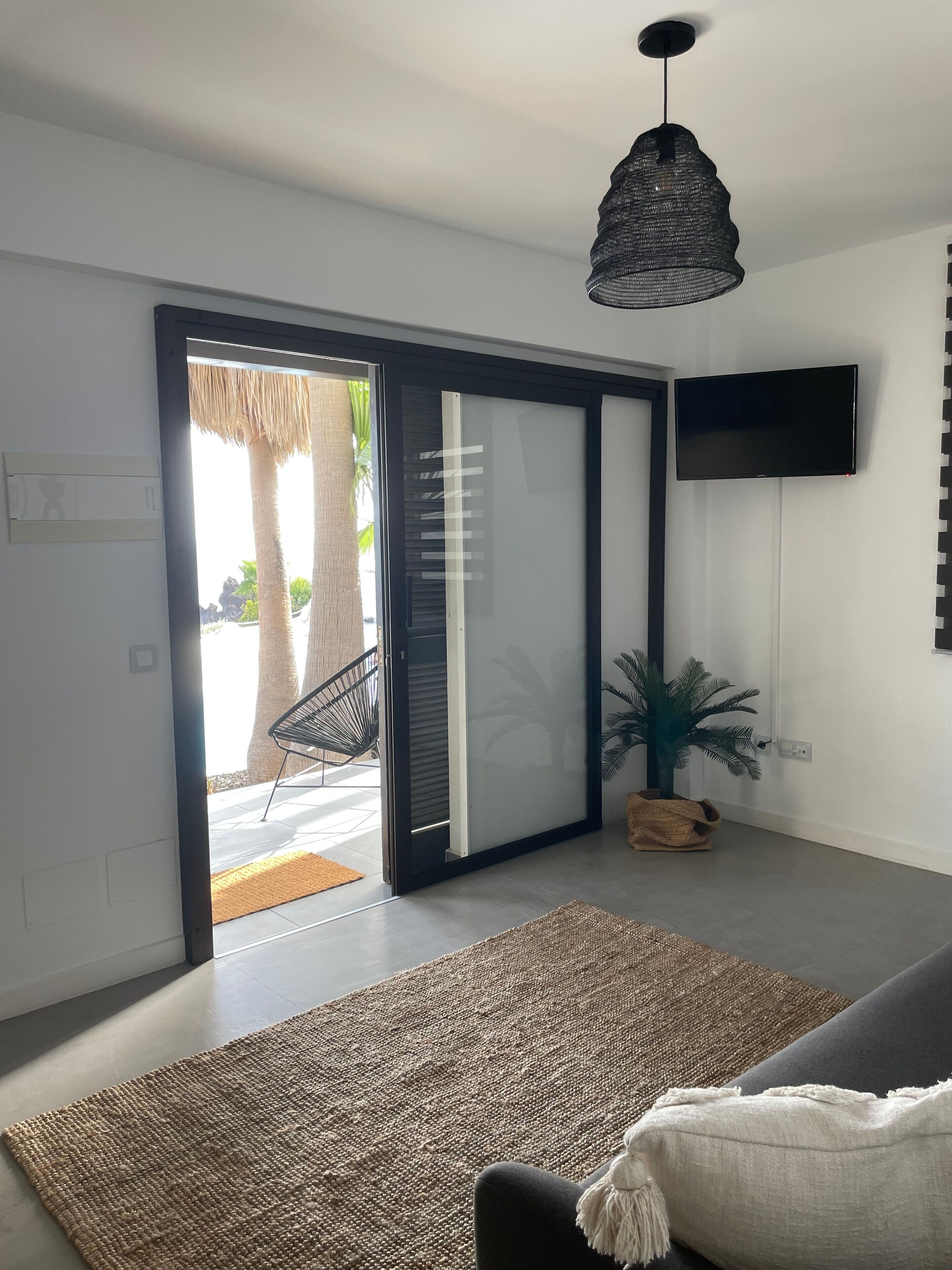 A modern living room with a sliding glass door leading to a balcony, a black pendant light, a flat-screen TV mounted on the wall, and a woven rug on the floor.