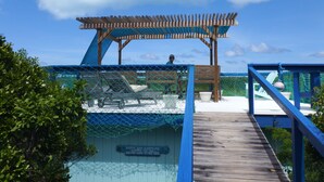 Eine Holzterrasse führt zu einem Strandbereich mit einer Pergola und Liegestühlen.