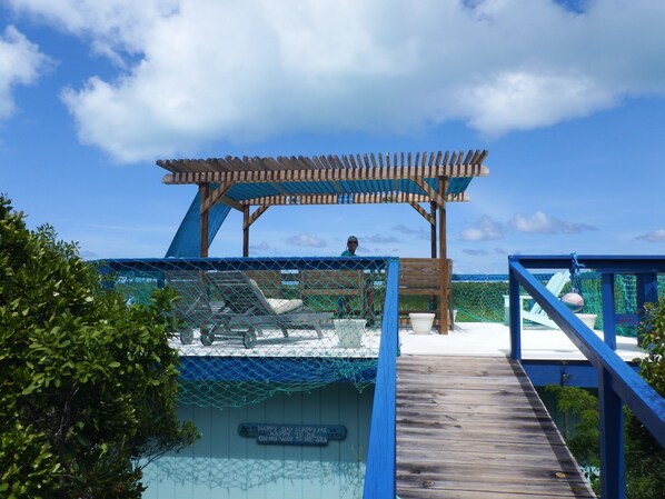 A wooden deck leading to a beach area with a pergola and lounge chairs.