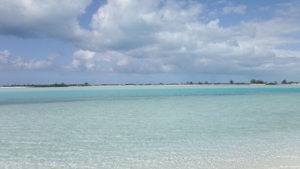 Ein Strand mit klarem türkisblauem Wasser und einer entfernten Küstenlinie mit spärlicher Vegetation.