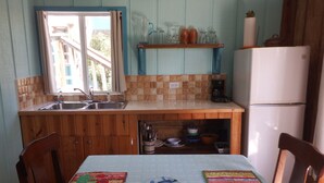 Eine kleine Küche mit Holzausstattung, Spüle und Kühlschrank. Ein Fenster mit Vorhängen, ein Regal mit Gläsern und ein Tisch mit Unterlagen.