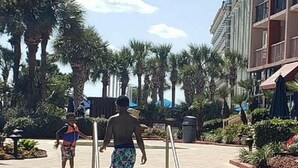 A swimming pool area with palm trees and a building in the background. - Beautifully Updated Condo in Beachfront Resort.   (Myrtle Beach)