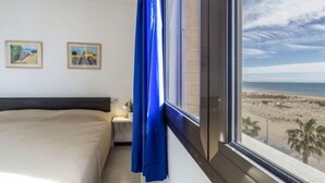 Ein Schlafzimmer mit einem großen Bett, einem Nachttisch, einem Fenster mit Blick auf den Strand und zwei gerahmten Bildern an der Wand.