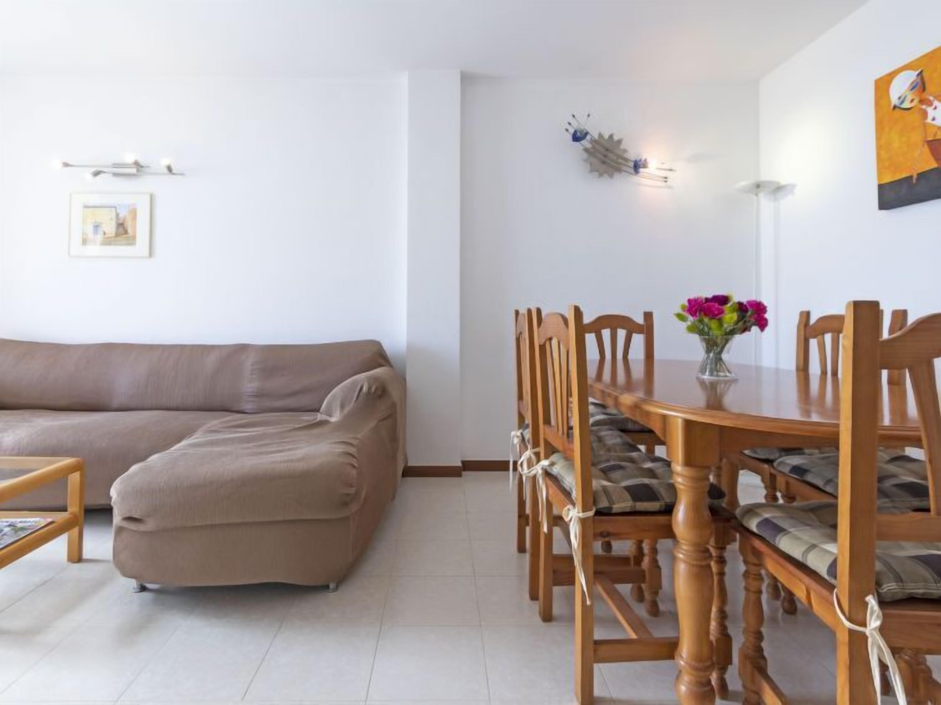 Una sala de estar con un sofá color beige, una mesa de comedor de madera con sillas, un jarrón con flores y una lámpara fijada a la pared.