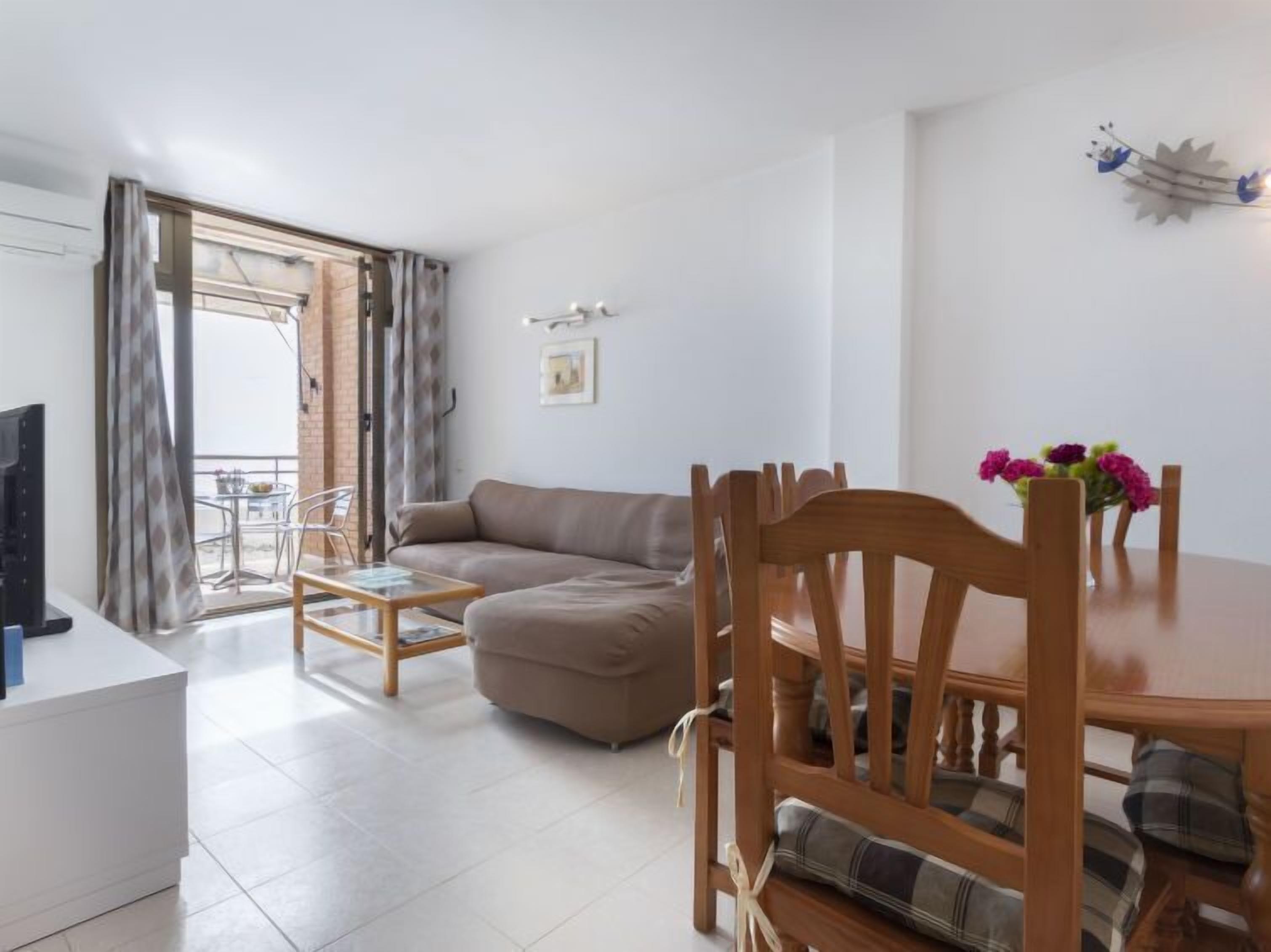 Ein modernes Wohnzimmer mit einer Couch, einem Holzesstisch und Stühlen. Ein Fernseher steht auf einer weißen Kommode, und es gibt einen Balkon mit Tisch und Stühlen.