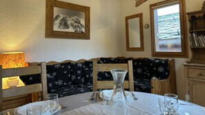 A dining area with a wooden table set for four, a bench with a snowflake pattern, a mountain painting, and a window with a view of a roof.