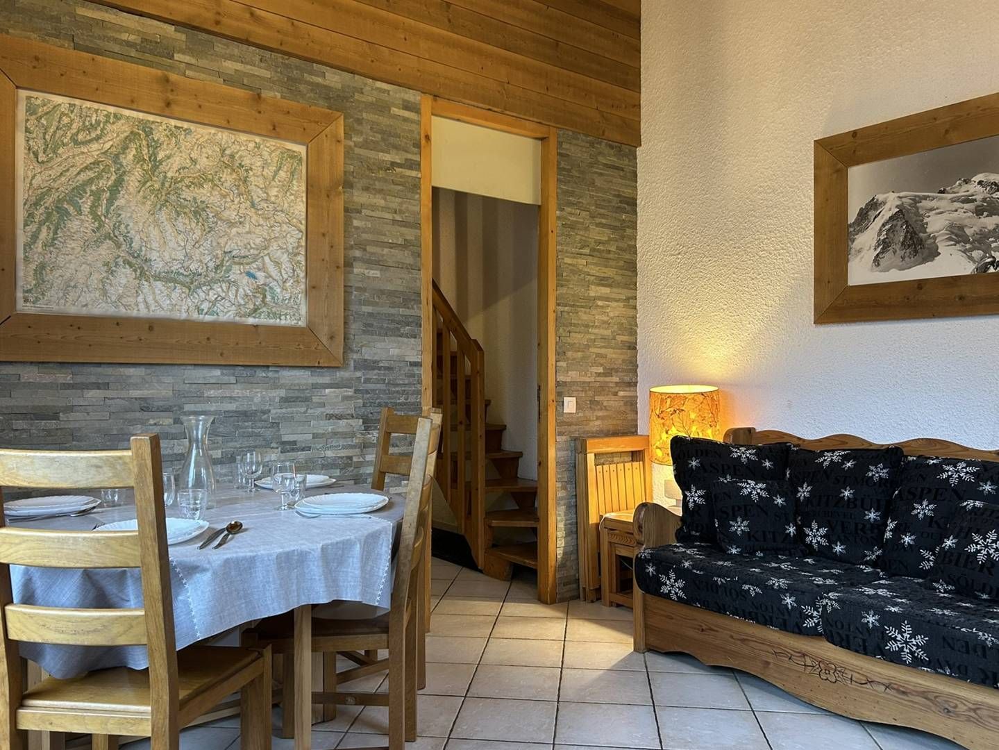 A dining area with a table set for two, wooden chairs, a map on the wall, and a sofa with a patterned cover.