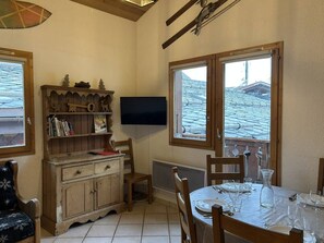 A dining area with a table set for two, a wooden cabinet with books and decorative items, and a television mounted on the wall.