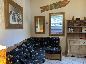 Een gezellige kamer met een houten bank, een klein kastje met boeken, een ingelijst beeld en een raam met uitzicht op een besneeerd landschap.
