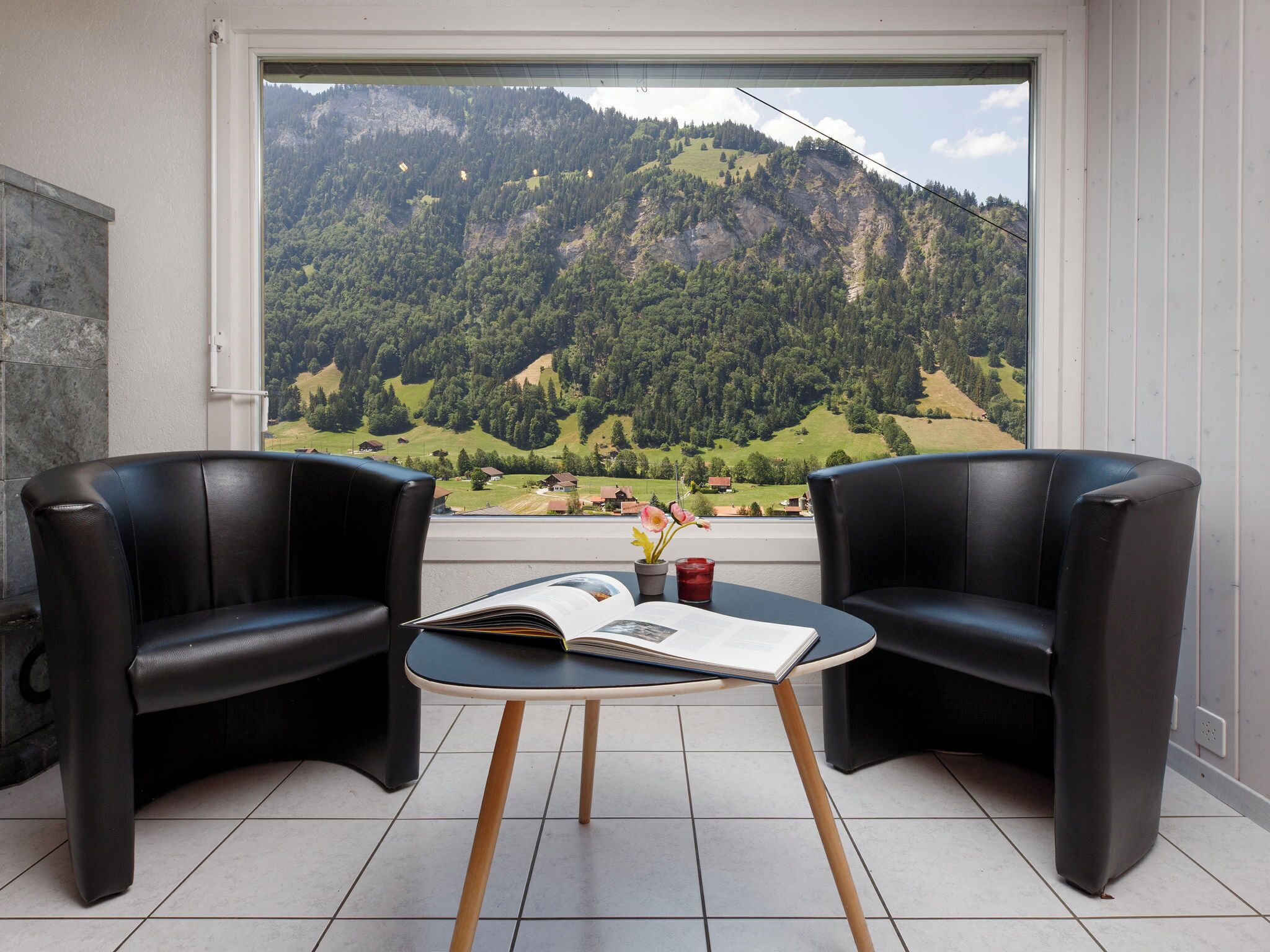 A room with two black leather chairs, a small table with a book and a glass of red drink, and a window with a mountain view.