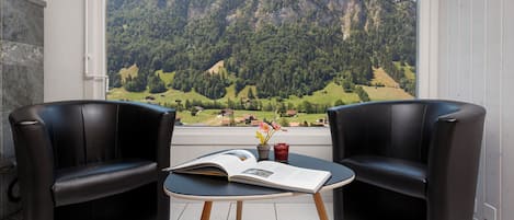 A room with two black leather chairs, a small table with a book and a glass of red drink, and a window with a mountain view.