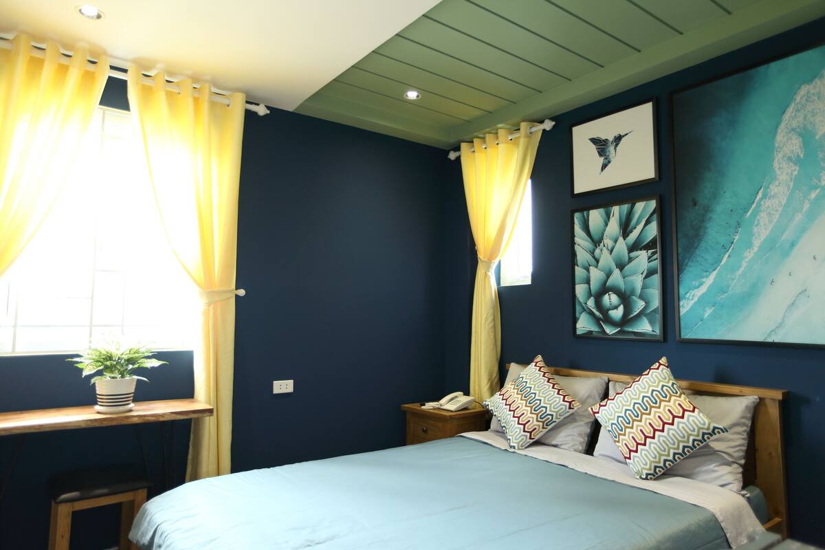 A bedroom with a bed, a bedside table, a nightstand, a window with curtains, and wall art.