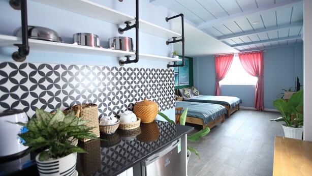 A modern kitchen with a black countertop, white and black patterned backsplash, and a stainless steel refrigerator. There are shelves above the counter holding pots and a potted plant. - Sleepbox Hostel Phu Quoc (Phu Quoc)