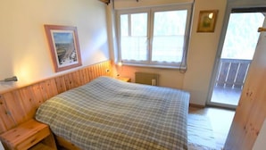 Une chambre à coucher avec un lit en bois, une table de chevet, une fenêtre avec des stores et une porte donnant sur un balcon.