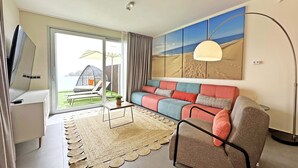 Een moderne woonkamer met een bank, een televisie, een vloerlamp en een groot raam met uitzicht op een strand.