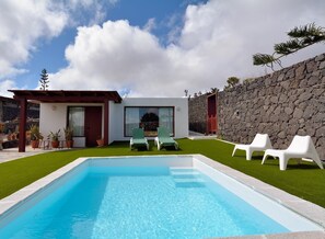 Ein moderner Poolbereich im Freien mit Steinmauer, einem kleinen Gebäude mit Glastür und zwei Liegestühlen.