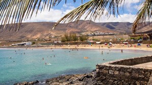 Une plage avec une eau turquoise et limpide, des gens qui se baignent et prennent des bains de soleil, et un mur de pierre au premier plan.