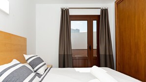 Une chambre d’hôtel avec une tête de lit en bois, un lit avec des oreillers à rayures, un couvre-lit blanc, une serviette blanche roulée et une vue à travers une porte avec des rideaux.