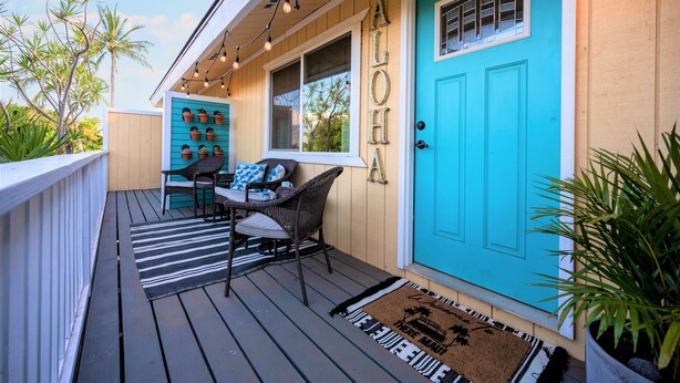 A porch with a turquoise door, a small seating area with wicker chairs, and a row of potted plants.