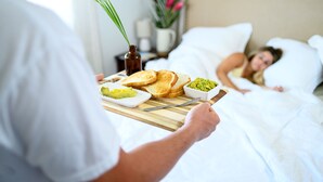 A breakfast tray with toast and avocado on a wooden board, held by a person in a white shirt.