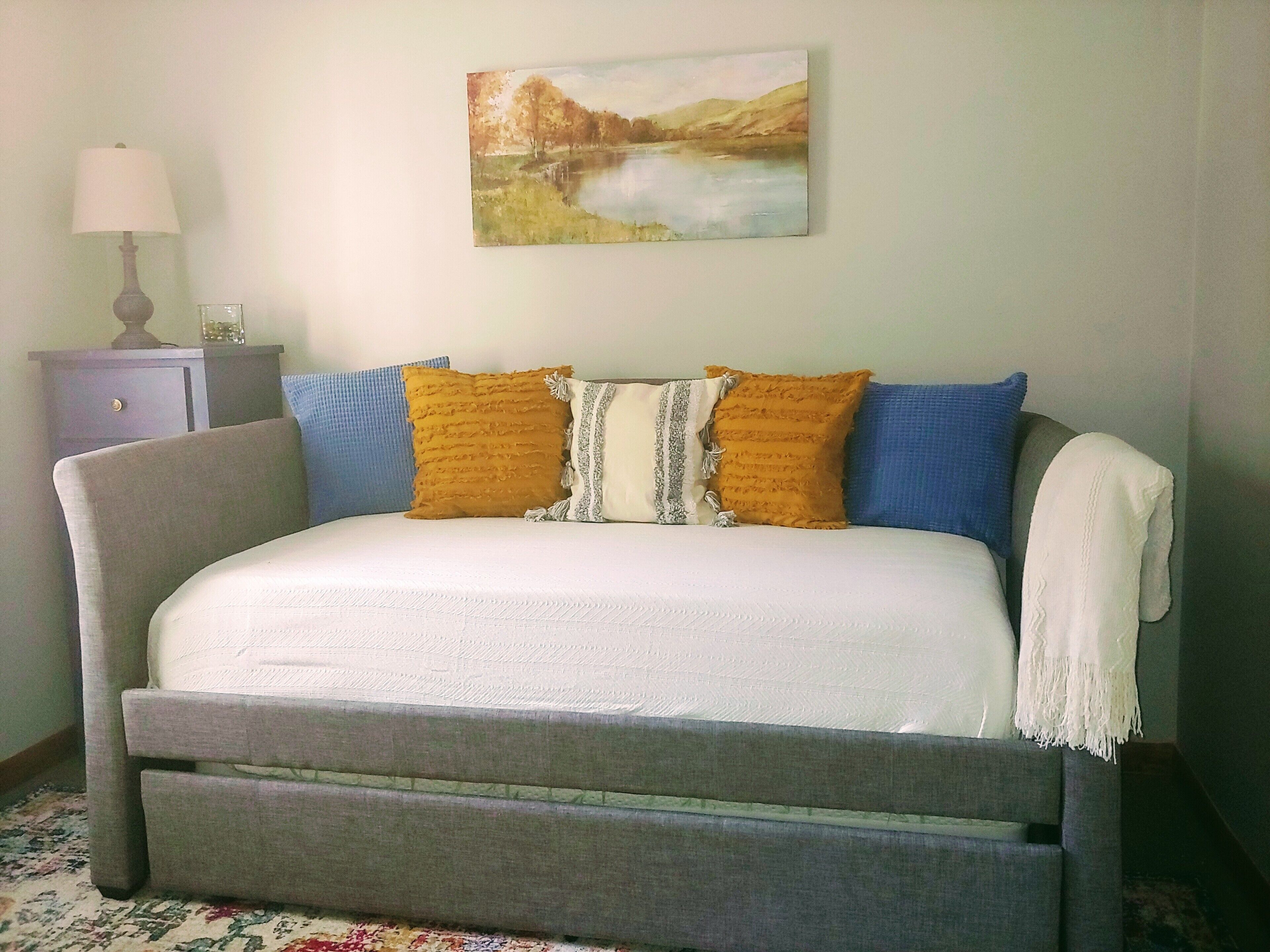 Una cama con colchón blanco, cabecera gris y almohadas azules y mostaza. Una lámpara y una mesita con un florero están sobre un aparador junto a la cama.