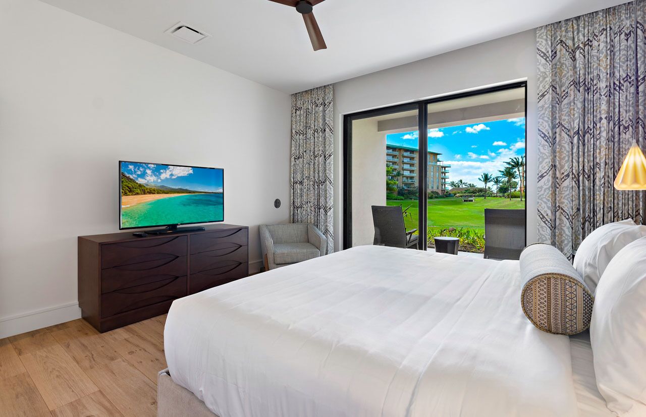 Ein Schlafzimmer mit einem großen Bett, einem Fernseher, einem Sessel und Blick auf eine grüne Landschaft durch eine Schiebetür.