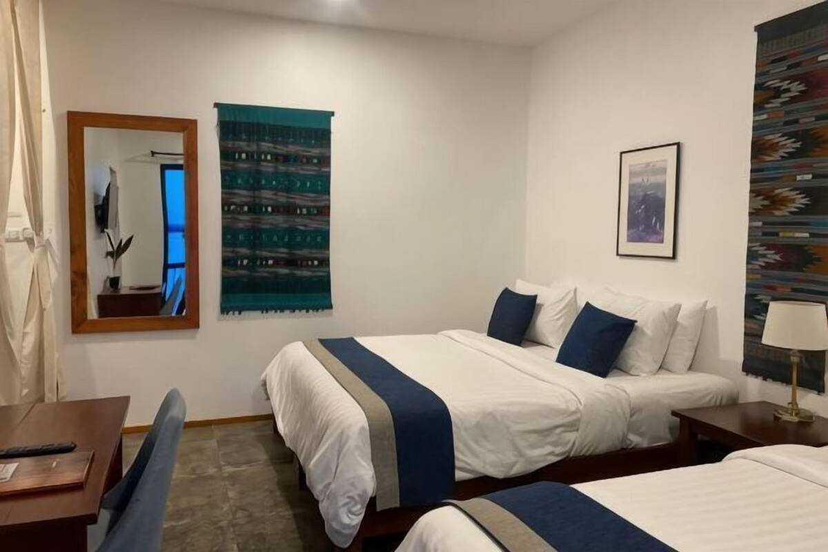 Habitación de hotel con dos camas, un escritorio, una silla, un espejo, un tapiz en la pared y un cuadro enmarco.