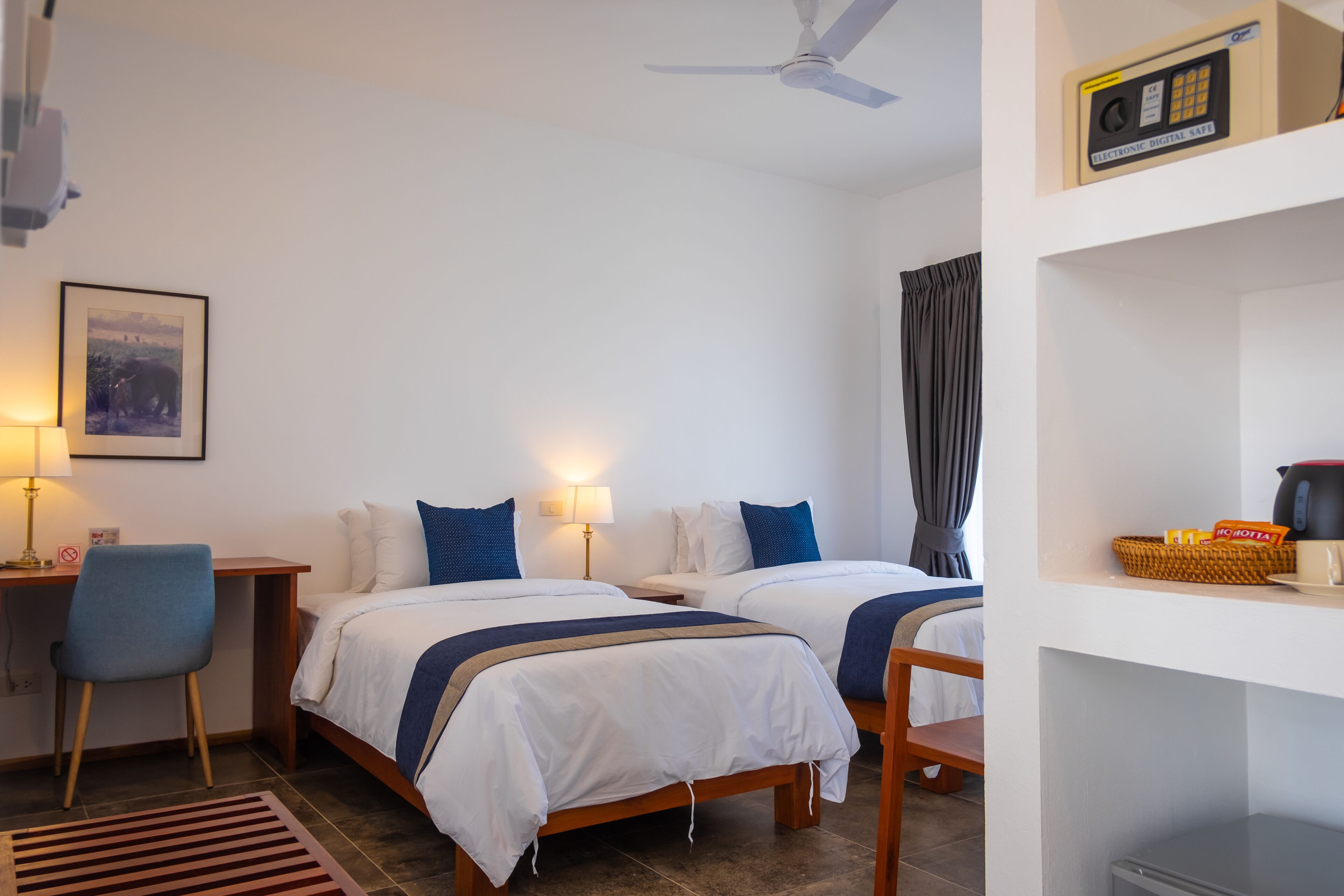 A hotel room with two beds, a desk, a chair, a ceiling fan, a framed picture on the wall, and a shelf with a kettle and a basket.