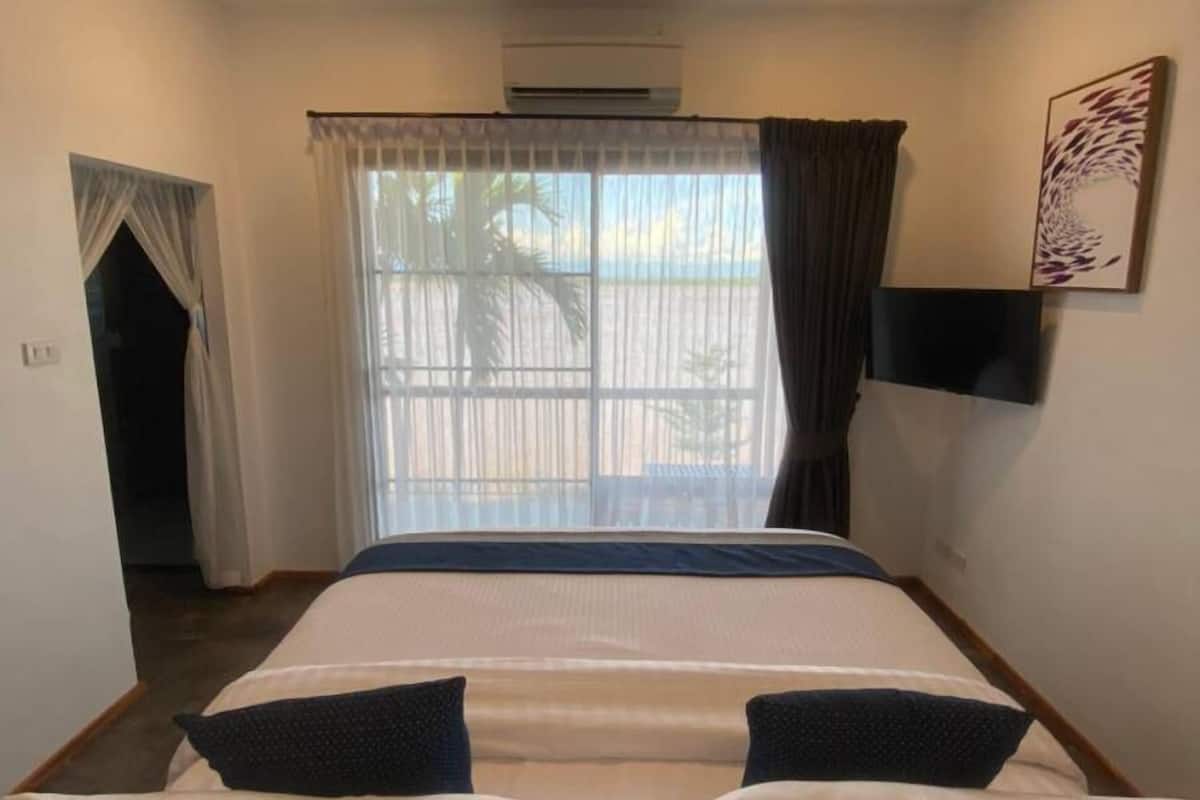 Habitación de hotel con cama, ventilador de techo, unidad de aire acondicionado, televisión y ventana con cortinas.