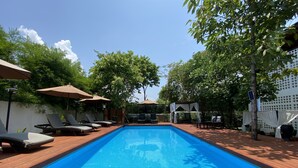 A rectangular swimming pool with a wooden deck surrounding it, shaded by umbrellas and trees.
