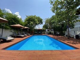 Una piscina rectangular con un deck de madera que la rodea, con sombrillas y árboles que brindan sombra.