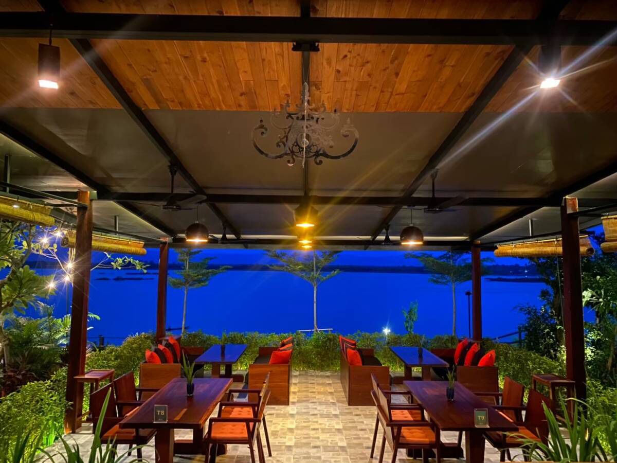 An outdoor dining area with wooden tables and chairs, a chandelier, and a view of the ocean at dusk.