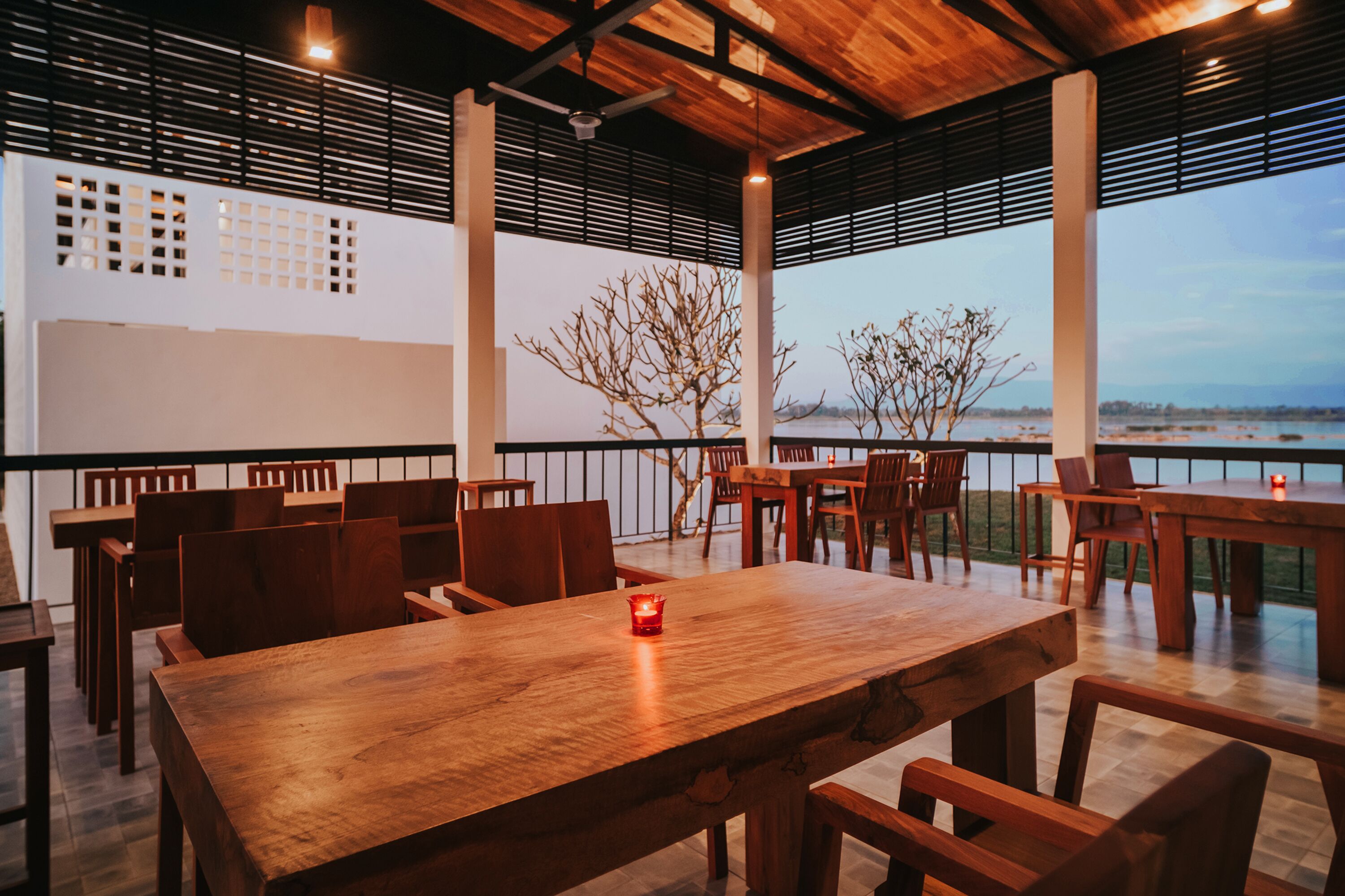 A restaurant with wooden tables and chairs, a candle on one table, and a view of the water.