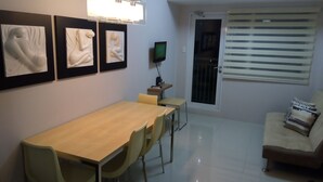 A dining area with a long table and chairs, a television on a stand, and a sofa with patterned cushions. - Grass homes 2 bedrooms (Bago Bantay)