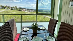 Un área de comedor con una mesa dispuesta para dos personas, una planta en maceta y una vista panorámica de una zona residencial a través de grandes ventanales.
