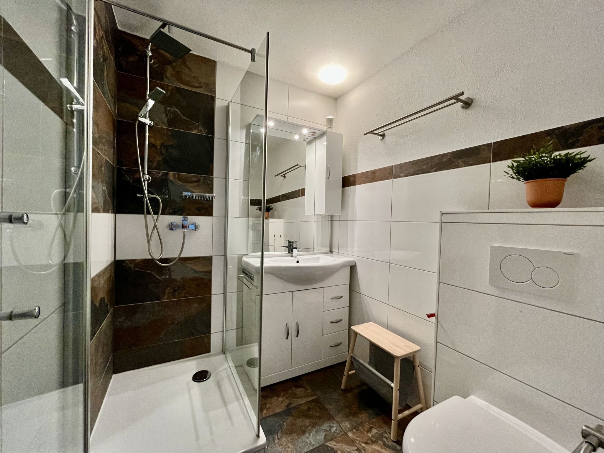 A modern bathroom with a glass-enclosed shower, a sink, a toilet, and a small wooden stool.