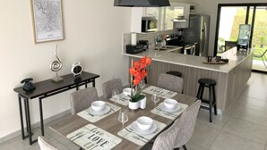 A modern kitchen with a central island, a dining area with a wooden table set for four, and a large window providing a view of the outdoors. - Exclusive Gated Community with HD TV, Air Conditioning, Cable and Netflix  (Nuevo Cuscatlán)