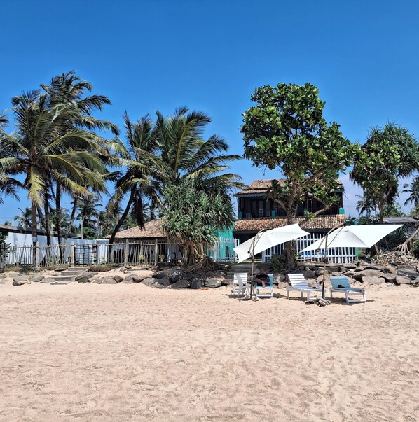 A beach with palm trees, a building with a tiled roof, and white umbrellas.