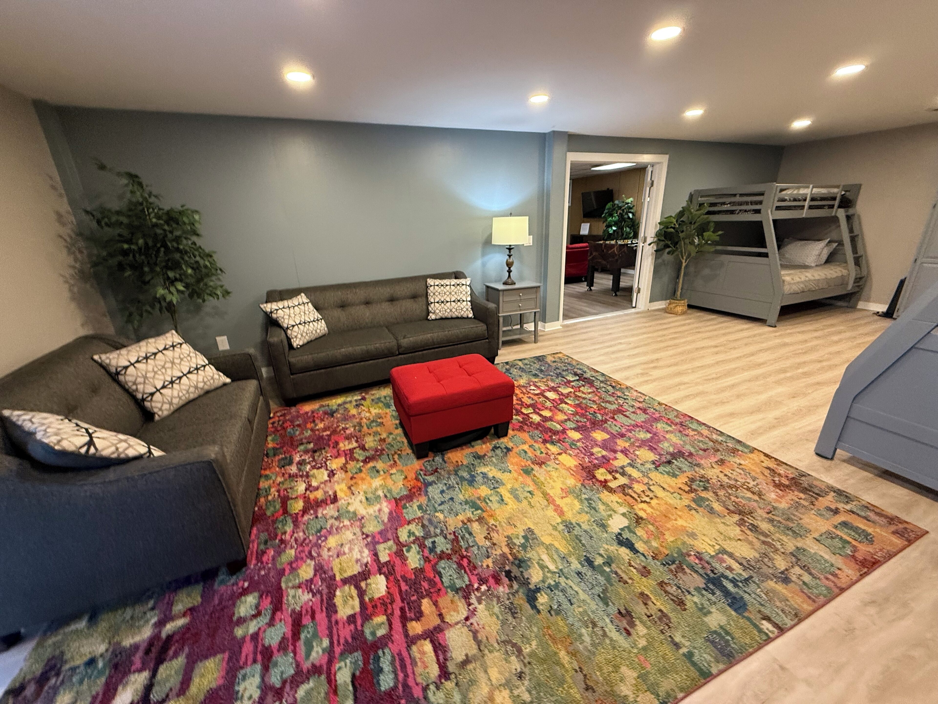 A living room with a sofa, ottoman, and colorful rug. There is a bunk bed in the background.