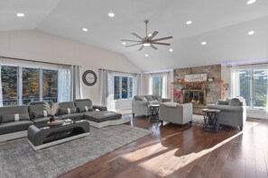 A spacious living room with a fireplace, large windows, and a ceiling fan.