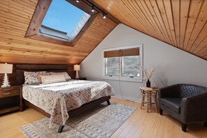 A bedroom with a sloped ceiling, a skylight, a bed with a floral bedspread, a dark leather armchair, and a small wooden table with a vase.